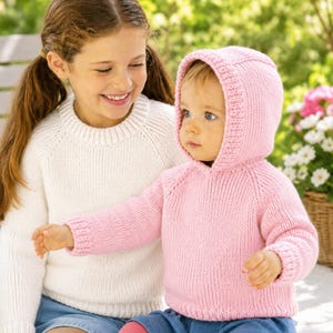 以下が含まれることがあります： 白いニットのセーターを着た若い女の子が、ピンクのフード付きセーターを着た赤ちゃんに微笑んでいます。赤ちゃんはまた、青いスカートとピンクのレギンスを着ています。背景は、緑と花のあるぼやけた屋外シーンです。