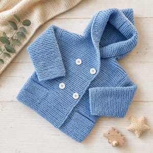 May include: A light blue knitted hooded jacket featuring a double-breasted front closure with four white buttons. The garment is crafted with a textured garter stitch pattern, long sleeves, and a hood, providing a consistent look throughout the piece.