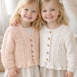 以下が含まれることがあります： お揃いのニットカーディガンを着た2人の若い女の子。1つはVネックの薄いピンク色、もう1つは丸首のクリーム色です。どちらも木製のボタンとテクスチャードニットのデザインです。
