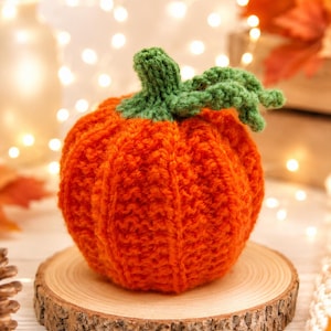 May include: A handmade orange crochet pumpkin with a green stem and leaves, displayed on a small wooden slice. This seasonal decoration is ideal for autumn or Thanksgiving. The background features soft, out-of-focus lights.