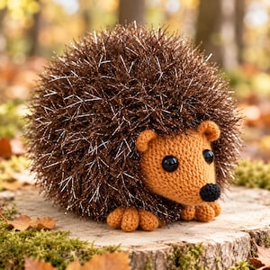 May include: A crocheted hedgehog plush toy featuring a round body covered in dark brown, shimmery, spiky yarn. The face, ears, and small feet are crafted from solid tan yarn, with two round black plastic eyes and a small black nose.