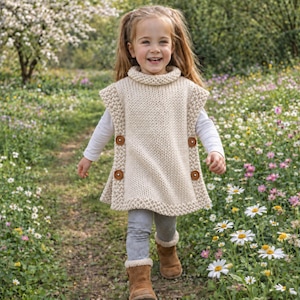May include: A cream-coloured, sleeveless, hand-knitted poncho with a high collar and a textured border. The garment features two brown wooden buttons on each side, securing the front and back panels together in a simple, functional design.