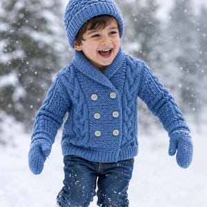 Könnte beinhalten: Ein Kind trägt eine blaue Strickmütze, einen Cardigan und Fäustlinge sowie eine blaue Jeans. Der Cardigan hat ein Zopfmuster und ist mit Knöpfen versehen. Das Kind lächelt und spielt im Schnee.
