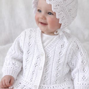 May include: A baby in a white knitted cardigan and bonnet. The cardigan features a V-neck, button fastening, and a scalloped edge. The bonnet has a matching scalloped trim and a ribbon tie. The baby is smiling.