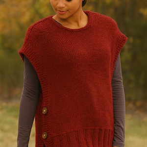 May include: A rust-colored knitted vest with a round neckline and short sleeves. The vest features two large, round wooden buttons on the side and a split hem. The model is wearing a long-sleeved gray shirt underneath.