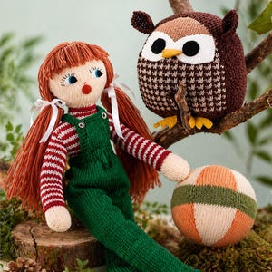 May include: A knitted doll with red hair, wearing a green overall and a red and white striped top. A knitted owl toy perches on a branch. A knitted ball with orange, green, and cream sections is nearby. The toys are on a mossy surface.