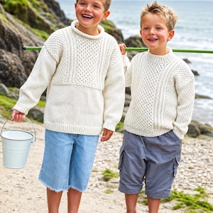 Op de afbeelding: Twee kinderen in crèmekleurige gebreide truien en spijkerbroekshorts staan op een strand. Eén houdt een kleine witte emmer vast. De truien hebben textuurpatronen. De achtergrond toont de zee en een rotsachtige heuvel.