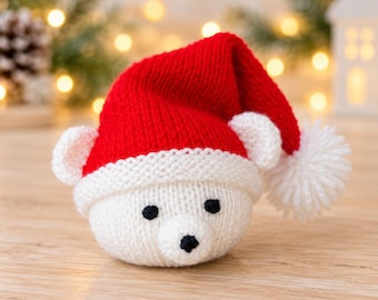 Puede incluir: Un adorno de cabeza de oso polar tejido en blanco que lleva un gorro de Papá Noel rojo brillante con borde blanco y un pompón blanco. El oso tiene dos orejas pequeñas blancas, dos ojos negros circulares y una nariz negra bordada.