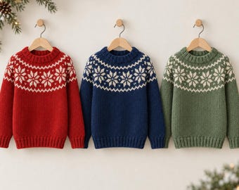 Puede incluir: Tres jerséis de punto de manga larga colgados en perchas de madera. Cada jersey presenta un cuello redondo y un patrón de copos de nieve blancos en el pecho y los hombros. Los jerséis son de color rojo sólido, azul marino y verde salvia.