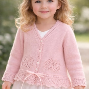 May include: A young child in a pale pink cardigan with a V-neck, button fastening, and butterfly-shaped cutouts. The cardigan has a scalloped hem and is tied with a ribbon. The child is also wearing a white dress.