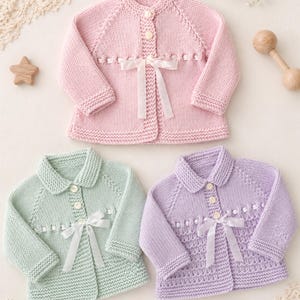 Pode incluir: Três cardigans de bebé de malha feitos à mão em cores pastel. O cardigan rosa tem gola redonda e é adornado com um laço de fita branco. Os cardigans verde e roxo têm gola Peter Pan e um laço de fita branco. Cada cardigan tem fechos de botão.