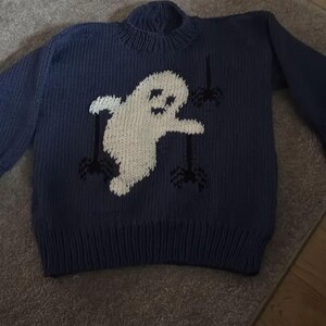 May include: A dark blue long-sleeve knit sweater featuring a large white ghost graphic centered on the front. The ghost is depicted with black eyes and a mouth, surrounded by three small black spider designs hanging from thin black lines.