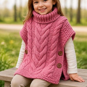 Pode incluir: Um suéter de malha rosa sem mangas com gola alta grossa, padrões de trança verticais na frente e botões castanhos ao longo das costuras laterais. A peça é usada sobre uma camisa branca de manga comprida e calças de cor clara.