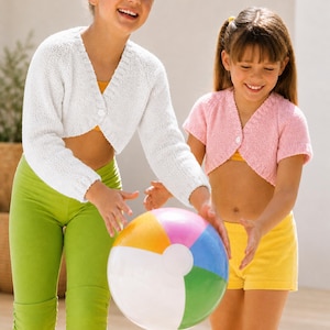 May include: Two young people are playing with a colorful beach ball. One is wearing a white, long-sleeved, cropped cardigan and green capri pants. The other is wearing a pink, short-sleeved, cropped cardigan and yellow shorts.