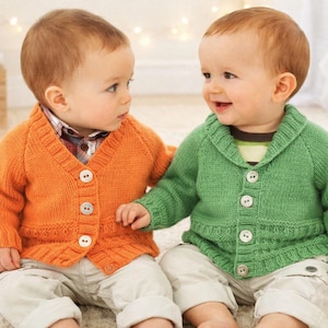 May include: Two babies wearing knitted cardigans. One baby is wearing an orange cardigan with white buttons, and the other is wearing a green cardigan with white buttons. Both babies are wearing light-coloured trousers.