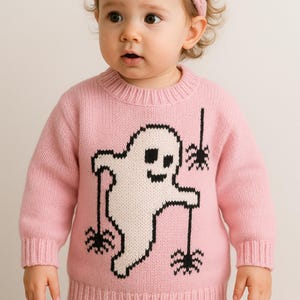 May include: A light pink long-sleeve knit sweater featuring a large white ghost graphic with black outlines. The ghost is depicted holding three black spider icons hanging from thin black lines. The sweater has a ribbed crew neckline, cuffs, and hem.