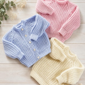May include: Three knitted jumpers in pastel colours: a light pink pullover, a light blue button-up cardigan with five small buttons, and a pale yellow pullover. Each garment features a textured, chunky knit pattern with long sleeves and ribbed cuffs.