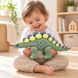 May include: A knitted dinosaur toy in sage green with a row of alternating lime green and grey circular plates along its back. The toy features a small red nose, embroidered black eyes, and a series of black triangular markings on its side.