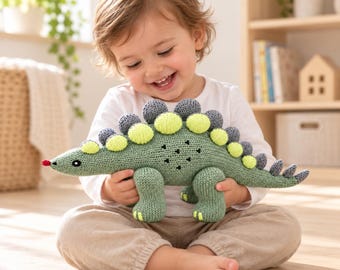 Puede incluir: Un juguete de dinosaurio tejido en color verde salvia con una hilera de placas circulares alternas en verde lima y gris a lo largo de su espalda. El juguete presenta una pequeña nariz roja, ojos negros bordados y marcas triangulares negras.