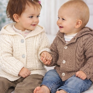 May include: Two babies wearing hooded cable-knit sweaters, one cream-colored and the other brown, sit close together. They are holding hands and smiling. The baby on the left wears brown pants, and the baby on the right wears blue jeans.