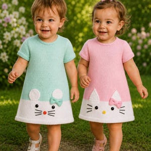 Puede incluir: Dos vestidos de punto de manga corta, uno en verde menta y otro en rosa claro, ambos con una sección inferior blanca con un diseño de cara de gato. Cada vestido incluye orejas de punto blanco, ojos de botón negros, bigotes bordados y un pequeño lazo.