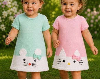 Puede incluir: Dos vestidos de punto de manga corta, uno en verde menta y otro en rosa claro, ambos con una sección inferior blanca con un diseño de cara de gato. Cada vestido incluye orejas de punto blanco, ojos de botón negros, bigotes bordados y un pequeño lazo.