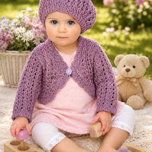 May include: A lavender knitted beret and a matching long-sleeve bolero cardigan featuring an openwork pattern and a single central button closure. The set is worn over a light pink dress and white leggings with eyelet lace trim at the cuffs.