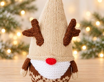 Puede incluir: Una figura de gnomo tejida con un gorro alto de color beige con astas marrones, una barba blanca, una nariz roja brillante y un cuerpo marrón con un patrón de chevrón beige en la base. El gnomo tiene pequeñas manos beige y brazos marrones.