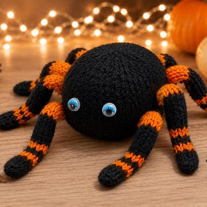 Puede incluir: Una araña negra tejida a mano con patas a rayas naranjas y negras y grandes ojos saltones. La araña es una decoración de Halloween, con calabazas y luces de hadas en el fondo.
