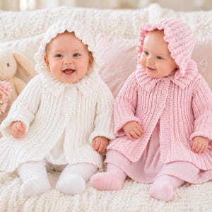 May include: Two babies wearing matching knitted outfits. One baby is in a white outfit with a bonnet and coat, and the other is in a pink outfit with a bonnet and coat. Both babies are smiling. A stuffed bunny and flowers are in the background.