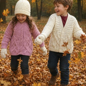 May include: Two children, a girl and a boy, are walking hand-in-hand through a park filled with autumn leaves. The girl wears a pink cable-knit cardigan, a cream-colored hat, and gloves. The boy wears a cream-colored cable-knit cardigan. Both wear dark blue jeans.