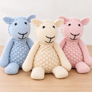 May include: Three stuffed sheep toys in light blue, cream, and light pink. Each sheep features a textured, popcorn-stitch body, smooth-knit head and limbs, and black embroidered eyes and mouth. The toys are seated upright.