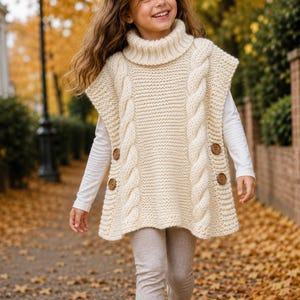 May include: A cream-colored sleeveless knit sweater vest featuring a chunky turtleneck, vertical cable knit patterns on the front, and two brown buttons on each side. The garment is worn over a long-sleeved white shirt and light-colored leggings.