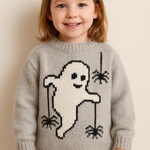 May include: A light gray long-sleeve knit sweater featuring a large white ghost graphic with black outlines. The ghost is depicted holding three black spiders hanging from thin black threads. The sweater has a ribbed crew neckline, cuffs, and hem.