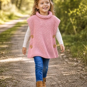 May include: A pink knitted poncho with a roll neck and large wooden buttons. The poncho is worn over a white long-sleeved top, paired with blue jeans and brown boots. The child is walking on a dirt path in a woodland setting.