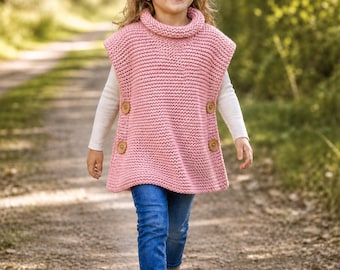 May include: A pink knitted poncho with a turtleneck collar and large wooden buttons. The poncho is worn over a white long-sleeved shirt and paired with blue jeans and brown boots. The child is walking on a dirt path.