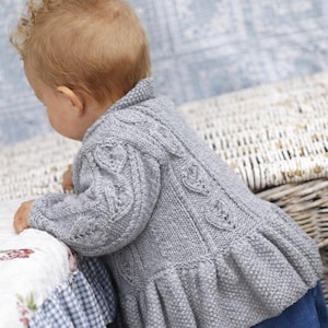 May include: A gray knitted baby sweater with leaf pattern details on the sleeves and back. The sweater has a ruffled hem and a collar. The baby is wearing blue jeans. The sweater is made of a soft, textured yarn.
