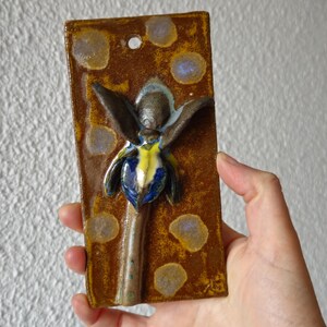 Può includere: Una placca da parete in ceramica con un fiore di iris stilizzato. La placca rettangolare marrone presenta dettagli marrone chiaro e blu. L'iris ha dettagli blu, gialli e marroni. Un foro nella parte superiore per appenderla.