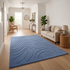Op de afbeelding: Een groot, rechthoekig blauw tapijt met een golvend patroon. Het tapijt ligt op een houten vloer en de kamer heeft een neutraal kleurenpalet met een bank, stoel en decoratieve elementen.