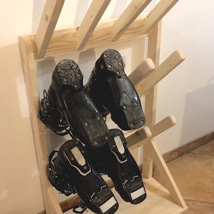 Può includere: Una rastrelliera in legno chiaro con supporti angolati progettata per riporre calzature. Quattro scarponi da sci neri sono montati sulla rastrelliera, con le suole rivolte verso l'esterno, mostrando i motivi del battistrada e le parti metalliche.