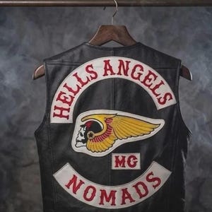 Puede incluir: Chaleco de cuero negro con parches bordados en blanco y rojo. Los parches dicen "HELLS ANGELS" y "NOMADS". También es visible un emblema de calavera alada. El chaleco está colgado de una percha de madera.