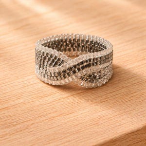 May include: A beaded ring with a woven design. The ring features clear and silver beads, creating a textured, intertwined pattern. The ring is set against a light wood surface.