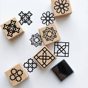 Può includere: Timbri in legno con disegni geometrici neri sono sparsi su una superficie bianca. I timbri sono quadrati e presentano vari motivi. È presente anche un tampone per inchiostro. I disegni stampati includono stelle, croci e altre forme.