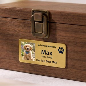 Pode incluir: Uma caixa de memorial de estimação de madeira com uma placa de identificação de latão. A placa de identificação apresenta uma foto de um filhote de golden retriever, o nome "Max", datas e a frase "Run free, Dear Max". A caixa tem um fecho de metal.