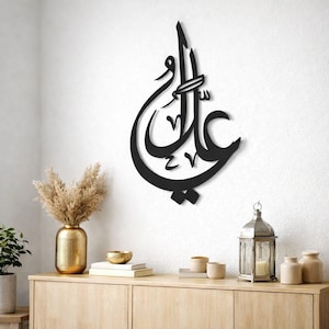 May include: Black metal wall art featuring Arabic calligraphy. The artwork is mounted on a white wall. The calligraphy is a stylized script with elegant curves and details. The wall art is a decorative piece.