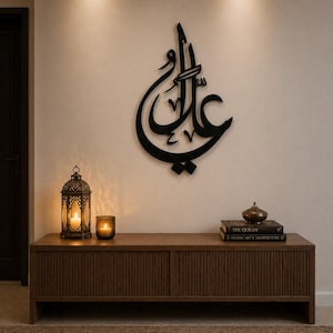 May include: Black metal calligraphy art piece in Arabic script mounted on a cream-colored wall. A wooden cabinet with a ribbed design holds a lantern, a candle, and books. The scene evokes a sense of cultural and spiritual decor.