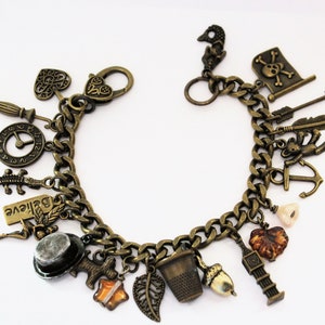 Könnte beinhalten: Ein goldfarbenes Charm-Armband mit einer Vielzahl von Charms, darunter eine Uhr, ein Anker, ein Herz, ein Fingerhut, eine Feder, ein Pfeil, ein Totenkopf mit Knochen, ein Hut, ein Blatt, eine Eichel, ein 'Glaube'-Schild und ein Krokodil.