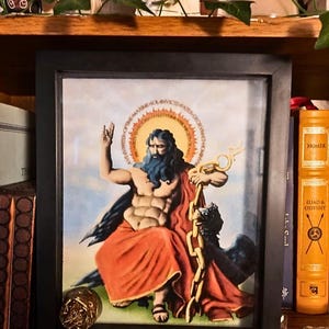 May include: Framed artwork featuring a seated figure with a beard, holding a golden chain and a spear. The figure is depicted with a halo and wearing a red robe. A black frame surrounds the artwork, with a gold coin in the foreground.