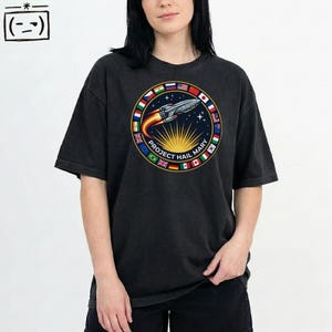 Può includere: T-shirt grigio scuro con un design circolare raffigurante un razzo, la scritta "PROJECT HAIL MARY" e le bandiere di diversi paesi. Il razzo ha fiamme e stelle su uno sfondo blu scuro. La maglietta ha maniche corte.
