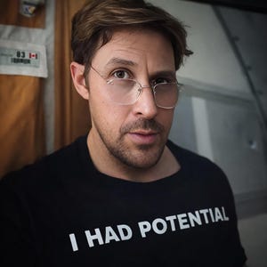 Pode incluir: Uma t-shirt preta com gola redonda com o texto branco "I HAD POTENTIAL". A pessoa está a usar óculos quadrados com armação fina. O fundo é de cor neutra. A imagem é um close-up do rosto e da parte superior do corpo.